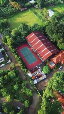 Etrafı yemyeşil ağaçlar ve evlerle çevrili bir tenis kortunun insansız hava aracı görüntüleri. Kırmızı çatılı bir bina sarayı kaplıyor..
