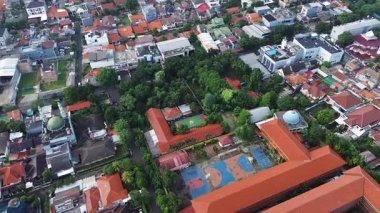 Yoğun yerleşim alanlarıyla çevrili bir okul kampüsünün insansız hava aracı görüntüleri. Kırmızı kiremit çatılar, yemyeşil alanlar ve basketbol sahaları görünür..