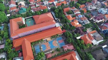 Kırmızı çatıları olan, yerleşim evleri ve yemyeşil ağaçlarla çevrili bir okul binasının insansız hava aracı görüntüleri. Basketbol ve diğer sahalar görülebilir.