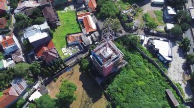 Evler ve yemyeşil yeşilliklerle çevrili, iletişim kulesi olan bir binanın yüksek açılı drone görüntüsü. Açık güneşli gün.