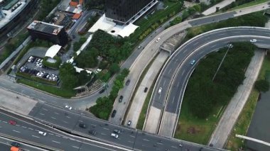 Otoyol kavşağının yüksek açılı görüntüsü, yoğun trafik akışını ve çevre şehir yapılarını gösteriyor. Yeşillik görünür.