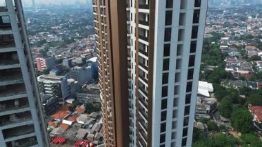 Yoğun bir şehrin ortasındaki modern bir apartmanın yüksek açılı görüntüsü. Çevredeki binalar ve yemyeşil alanlar görülebilir..