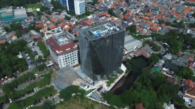Geleneksel evler ve verimli yeşilliklerle çevrili modern bir gökdelenin yüksek açılı görüntüsü. Binanın yanında bir kanal var..