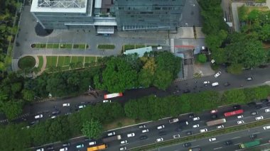 Modern bir ofis binasının yanındaki yoğun şehir trafiğinin insansız hava aracı görüntüleri. Yol boyunca yemyeşil ağaçlar var. DKI Jakarta, Endonezya - 22 Ocak 2025