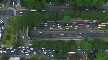 Yoğun trafik sıkışıklığı olan yoğun şehir otoyolu yukarıdan görüldü. Araçlar yavaş hareket ediyor. Yol boyunca yemyeşil ağaçlar var..