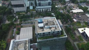 Kalabalık bir şehirde, otoparklar ve yemyeşil alanlarla çevrili modern bir ofis binasının insansız hava aracı görüntüleri. DKI Jakarta, Endonezya - 22 Ocak 2025