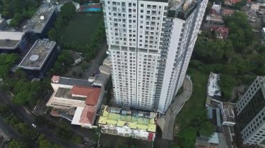 Şehir evleri ve yollarla çevrili modern bir apartmanın yüksek açılı insansız hava aracı görüntüsü. Yemyeşil ağaçlar.