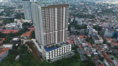 Yoğun bir şehir manzarası içinde, çeşitli binalar ve yemyeşil alanlarla çevrili modern bir apartmanın yüksek açılı görüntüsü..