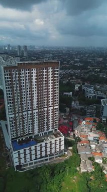 Bulutlu bir gökyüzünün altındaki yoğun şehir manzarasının ortasındaki uzun ve modern bir apartmanın insansız hava aracı görüntüleri. Etrafını saran evler ve şehrin ufuk çizgisi görünür.