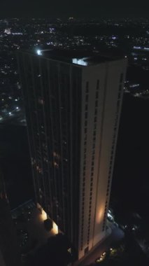 Gece modern bir gökdelenin yüksek açılı görüntüsü, karanlık bir şehrin arka planına karşı aydınlatılmış. Şehrin çevresindeki ışıklar görünür..