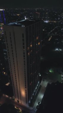 Şehir ışıkları ve koyu yeşilliklerle çevrili modern bir gökdelenin yüksek açılı görüntüsü..