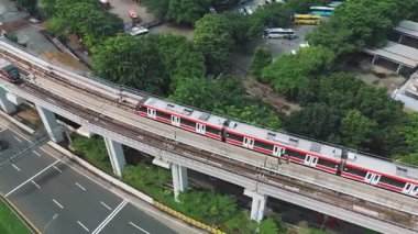 Yeşillik ve kentsel yayılma ile çevrili bir otobanın yakınındaki yüksek bir raydaki trenin hava görüntüleri. DKI Jakarta, Endonezya - 01 Şubat 2025