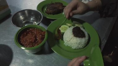 Bir tabak pilava sambal ekleyen ellerin yukarıdan çekimi. Pirinç, salatalık ve ızgara ördek görülebilir..