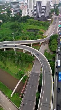 Karayolu trafiği, üstgeçit, şehir binaları ve yemyeşil alan. Araçlar birden fazla şerit boyunca hareket eder.