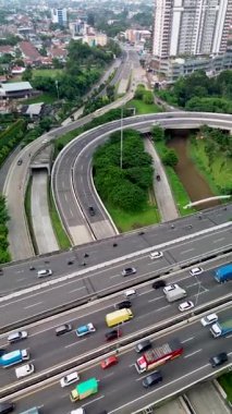 Otoyol trafiğinin yüksek açılı görüntüsü, hareket halindeki araçlar, şehir binaları ve görünür yeşil alanlar.