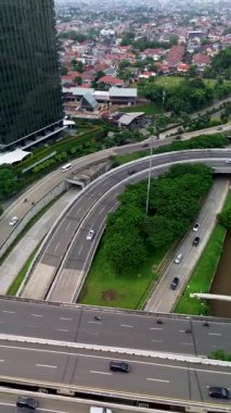 Otoyol trafiği, şehir binaları ve yeşil alanlar yüksek açılı. Arabalar, kamyonlar ve motosikletler birden fazla şerit boyunca hareket ederler..