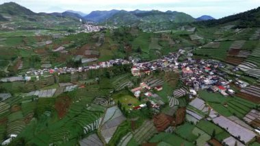 Bu hava görüntülerinde teraslı pirinç tarlaları, evleri olan küçük bir köy ve bulutlu bir gökyüzünün altındaki uzak dağlar yer alıyor. Kamera yavaşça sola döner..