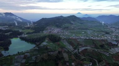 İnsansız hava aracı görüntüsü Endonezya 'daki Dieng Platosu' nu gösteriyor. Bir göl, teraslı tarım arazileri, küçük bir kasaba ve puslu bir gökyüzünün altındaki uzak dağlar..