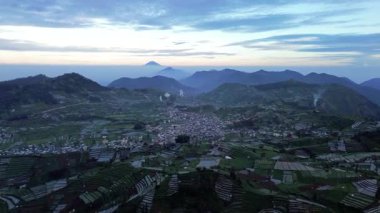 Bu hava görüntülerinde, yemyeşil tarlalarla çevrili bir vadide yuva yapmış bir kasaba görülüyor. Dağlar arka planda yükselir yumuşak bir gündoğumu gökyüzünün altında.