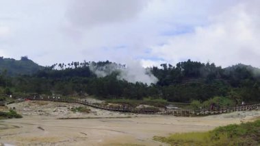 Görüntüler yerden buhar sızıntısı olan bir jeotermal alan gösteriyor. Ahşap bir geçit, yürüyen insanlar ve ağaçlı yeşil bir yamaç var. Bulutlu gökyüzü.