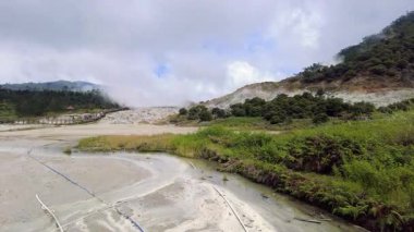 Görüntülerde Sikidang Krateri 'nin tahta köprüsü, yeşil bitki örtüsü ve bulutlu bir gökyüzü var. Yamaçtaki kraterden buhar yükseliyor..