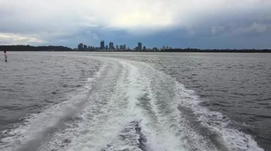 Kuğu Nehri 'nde seyreden bir teknenin bıraktığı köpüklü su izinin yavaş çekim görüntüsü, Perth şehrinin ufuk çizgisi uzakta..