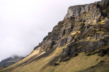 Bu görkemli kayalık duvar, bitki örtüsü tarafından hafifçe yumuşatılmış dikey uçurum ve uçurumların karmaşık bir dokusuna sahiptir. Bulutlu gökyüzü bu vahşi doğaya karamsar, atmosferik bir his katıyor. İzlanda