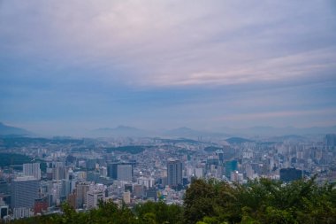 Seul şehri ufka doğru yayılıyor, yumuşak bulutlu gökyüzünün altında yuvarlanan dağlar tarafından destekleniyor..