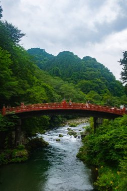 Nikko, Japonya - 12 Ağustos 2024: Shinkyo Köprüsü, yemyeşil orman ve tepelerle çevrili, açık hava meraklıları ve kültürel keşifler için ideal sakin ve pitoresk bir doğal manzara tasvir eden, akan bir nehir üzerinde uzanır..