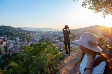 Saraybosna, Bosna. 25 Temmuz 2019 'da Bosna-Hersek' in başkenti Saraybosna 'daki Park Princeva Restoranı' nın terasından gün batımını izleyen insanlar görülüyor.