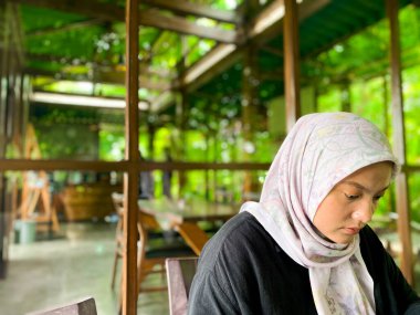 Çiçekli tesettürlü bir kadın yemyeşil bir kafede huzur içinde oturur. Atmosfer sakin ve sakin..