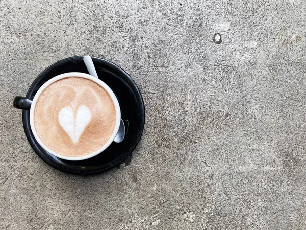 Kalp şeklinde bir latte sanat kremalı bir kahveyi süslüyor, kırsal beton bir masanın üstünde siyah bir fincanda servis ediliyor..