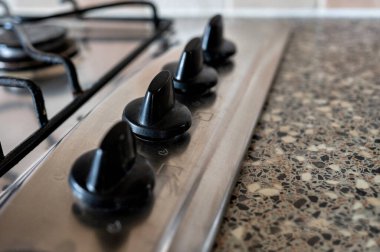 Mavi alev ve metal ızgaraların yer aldığı tutuşturulmuş bir gaz ocağının yakın çekimi fayanslı bir mutfak arka planına dayanır, mutfak atmosferini ve ev yemeklerini ön plana çıkarır