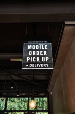 Bir kahve dükkanının içinde asılı bir tabela Mobil Sipariş Teslimatı. Fotoğraf modern iç mekanın bir kısmını tavan lambaları ve endüstriyel dekorla gösteriyor. Starbucks.