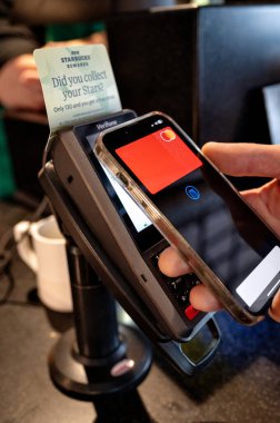 Bir kişi, bir kahve dükkanında ulaşılamaz bir ödeme yapmak için akıllı telefon kullanır. Ödeme terminali ve telefonu tutan elin bir kısmı görünür..
