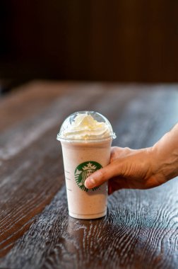 A hand holding a takeaway Starbucks frappuccino with whipped cream, placed on a wooden table inside a coffee shop in Corby, UK.