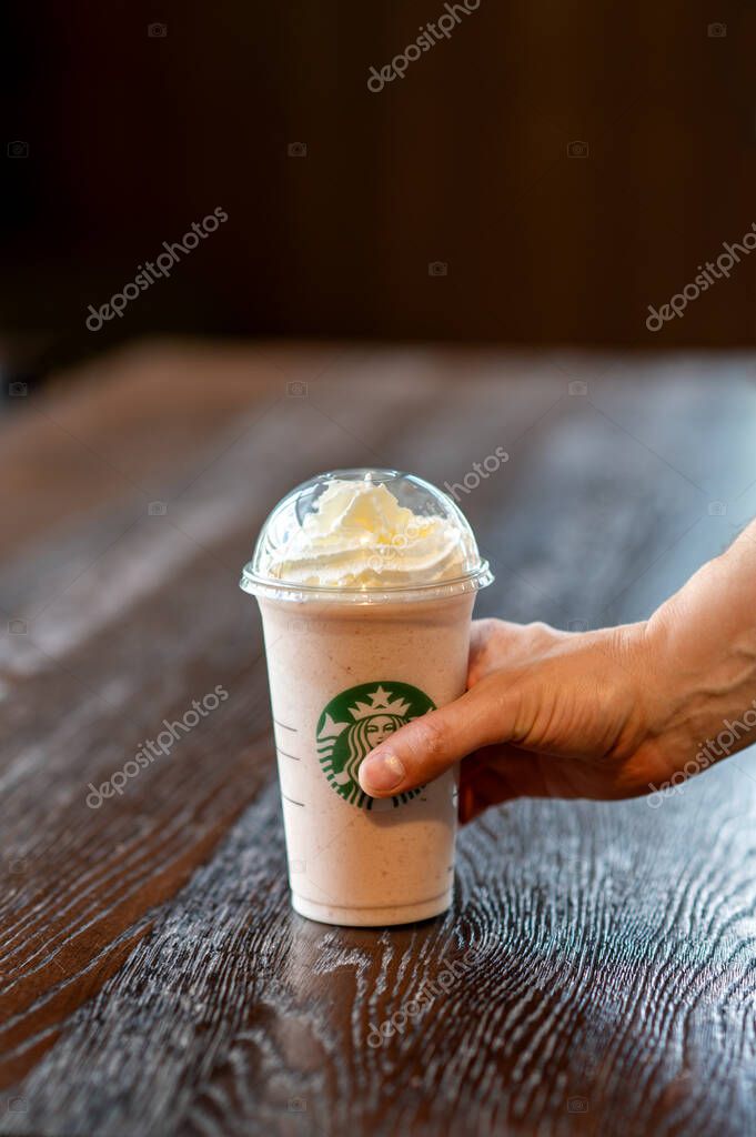 A hand holding a takeaway Starbucks frappuccino with whipped cream, placed on a wooden table inside a coffee shop in Corby, UK.