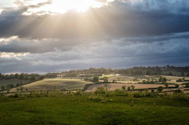 Altın güneş ışınları, karanlık bulutların arasından kırsal manzaralı tarlaları ve dağınık evleri aydınlatıyor. Umut verici ve ruhani bir atmosfer yaratmak..