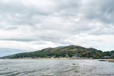 Kumlu bir sahil kenarına kurulmuş küçük bir sahil köyü ile denize doğru uzanan yeşil yamaçlı yarımadanın manzarası. 
