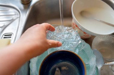 Bir el, cam kaseyi bir su deresinin altında duruluyor. Kirli tabaklar lavaboya istiflenir, kaseler, bardaklar ve çatal bıçaklar dahil. Yüksek kalite fotoğraf