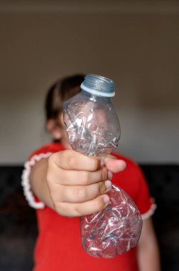 Bir çocuk, erken eğitim yoluyla geri dönüşümü, çevre sorumluluğunu ve atıkların azaltılmasını simgeleyen ezilmiş plastik bir şişeyi kameraya doğru tutuyor. Yüksek kalite fotoğraf