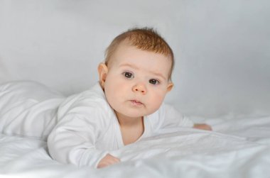 Beyaz elbiseli şirin bir bebek yumuşak bir yatakta yatıyor ve ileriye bakıyor..