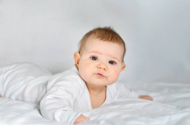 Beyaz elbiseli şirin bir bebek yumuşak bir yatakta yatıyor ve ileriye bakıyor..