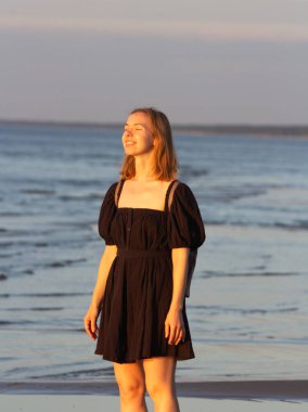 Genç bir kadın kumlu bir sahilde duruyor, canlı günbatımının tadını çıkarıyor. Kadın figürü, okyanusun kenarındaki renkli alacakaranlığı huzur içinde gözlemler. Huzurlu bir sahil sahnesi, huzurlu bir akşam. Yüksek kalite fotoğraf