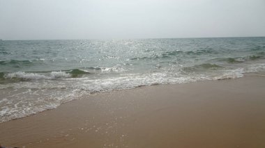 Yumuşak, puslu bir gökyüzünün altında kumlu kıyılarda dalgaların dalgalandığı huzurlu bir sahil sahnesi. Işık sudan yansıyarak, sakin atmosfere katkı sağlar..