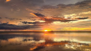 Yüzeyine yansıyan canlı turuncu, sarı ve kırmızı renkleri olan sakin bir su kütlesinin üzerinde nefes kesici bir günbatımı. Gökyüzü dramatik bulutlarla bezenmiş, resim gibi ve huzurlu bir sahne yaratıyor..