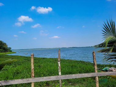 Bereketli bir yeşillik, sakin bir mavi göl ve berrak bir gökyüzü olan sakin bir göl manzarası. Tahta direkler ve palmiye yaprakları ön planda çerçevelenerek, kırsal bir dokunuş ekliyor.