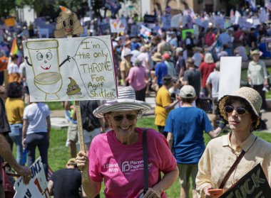 Karikatür tuvaleti ve kaka emojisi olan komik bir tabela tutan bir kadınla protesto sahnesi. Arka plandaki kalabalık, siyasi bir miting veya protesto anlamına gelen çeşitli işaretler taşıyor.