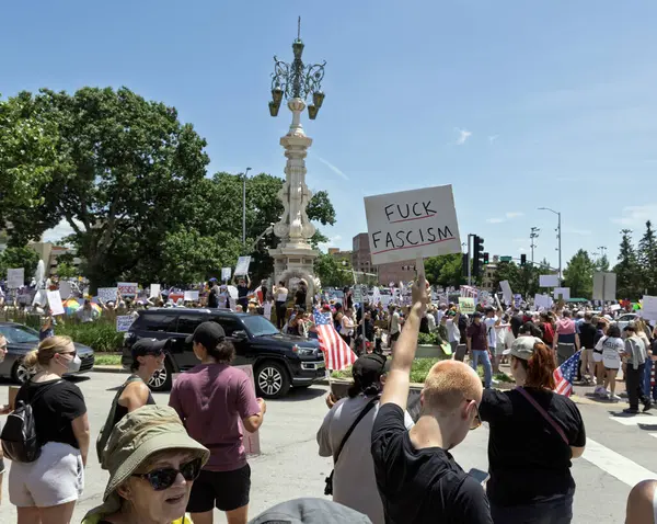 Büyük bir kalabalık, elinde siyasi mesajlarla bir şehir meydanında protesto için toplanıyor. Sahnede çeşitli katılımcılar, araçlar ve açık mavi gökyüzünün altında tarihi bir sokak lambası yer alıyor..