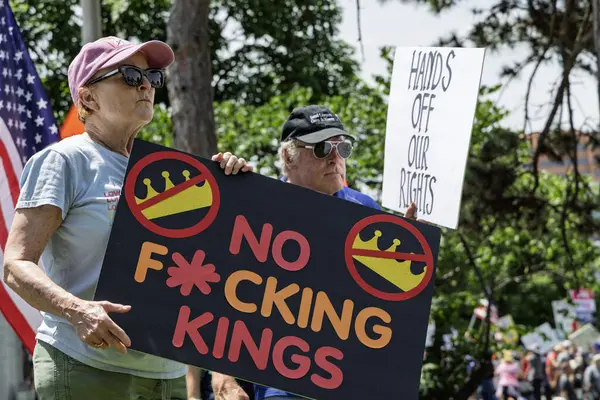 İki yaşlı protestocu bir mitingde tabela tutuyor. Bir tabelada 'F * cking Kings yok' yazıyor, arkasında da 'Eller Haklarımızdan Uzak' yazan bir tabela var. Ağaçlar ve bayraklar görünür.